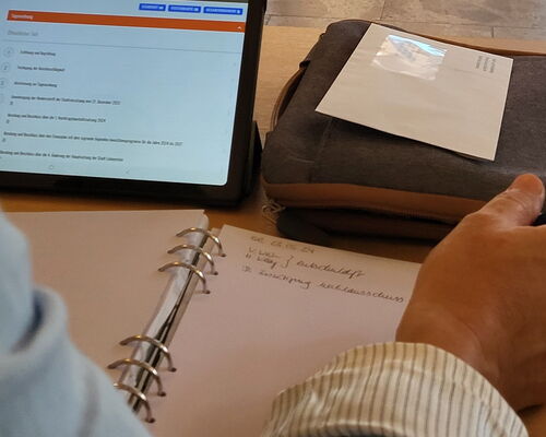 Tisch mit Tablet, auf dem eine Tagesordnung angedeutet ist, Notizblock und Person, die Stift in der Hand hält