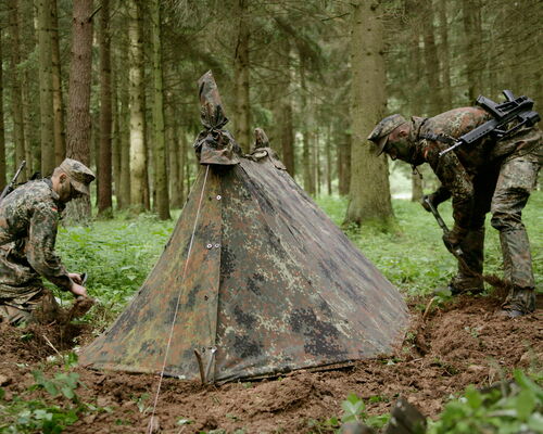2 Rekruten haben im Wald ein Zelt aufgebaut und graben eine Abflussrinne für Regenwasser