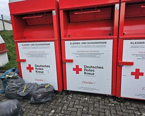 drei rote Altkleidercontainer mit dem roten Kreuz, davor graue riesige Müllsäcke