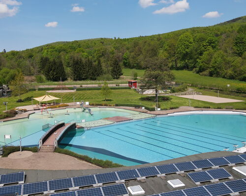 Luftaufnahme des Freibades mit Schwimmbecken, Nichtschwimmerbecken, Liegewiesen und Bergen im Hintergrund