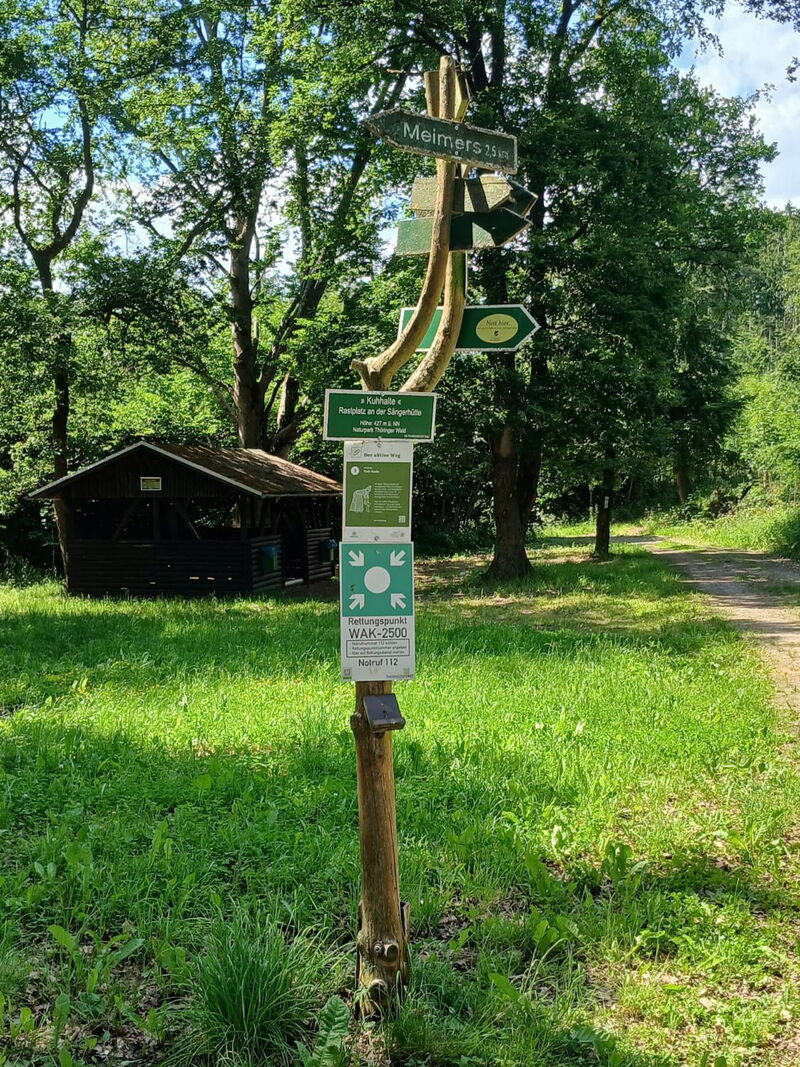 Wegweiser mit Ortsangabe Kuhhalte an der Sängerhütte, im Hintergrund ein Holzhaus am Waldrand