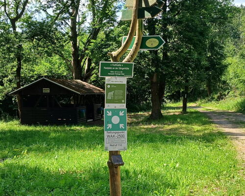 Wegweiser mit Ortsangabe Kuhhalte an der Sängerhütte, im Hintergrund ein Holzhaus am Waldrand