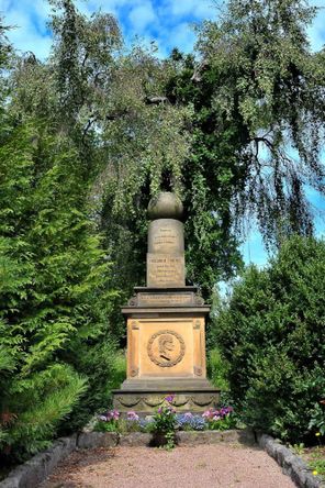 Grabdenkmal vor Bäumen und blauen Himmel. Denkmal besteht aus Kugel, Würfel, Walze, trägt Reliefbild von Friedrich Fröbel und Zitat "Kommt lasst uns unseren Kindern leben"