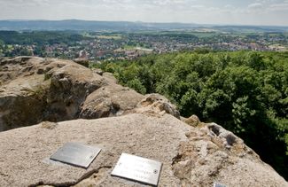 Panorama: Turmfelsen der Burgruine mit Richtungsplaketten im Vordergrund, rechts grüne Laubbäume, in der Mitte Dächerlandschaft von Bad Liebenstein, im Hintergrund Berge und blauer Himmel