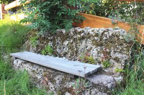 steinerne Bank aus einem Stück mit Holzauflage, bewachsen mit Moos und Steingartenpflanzen