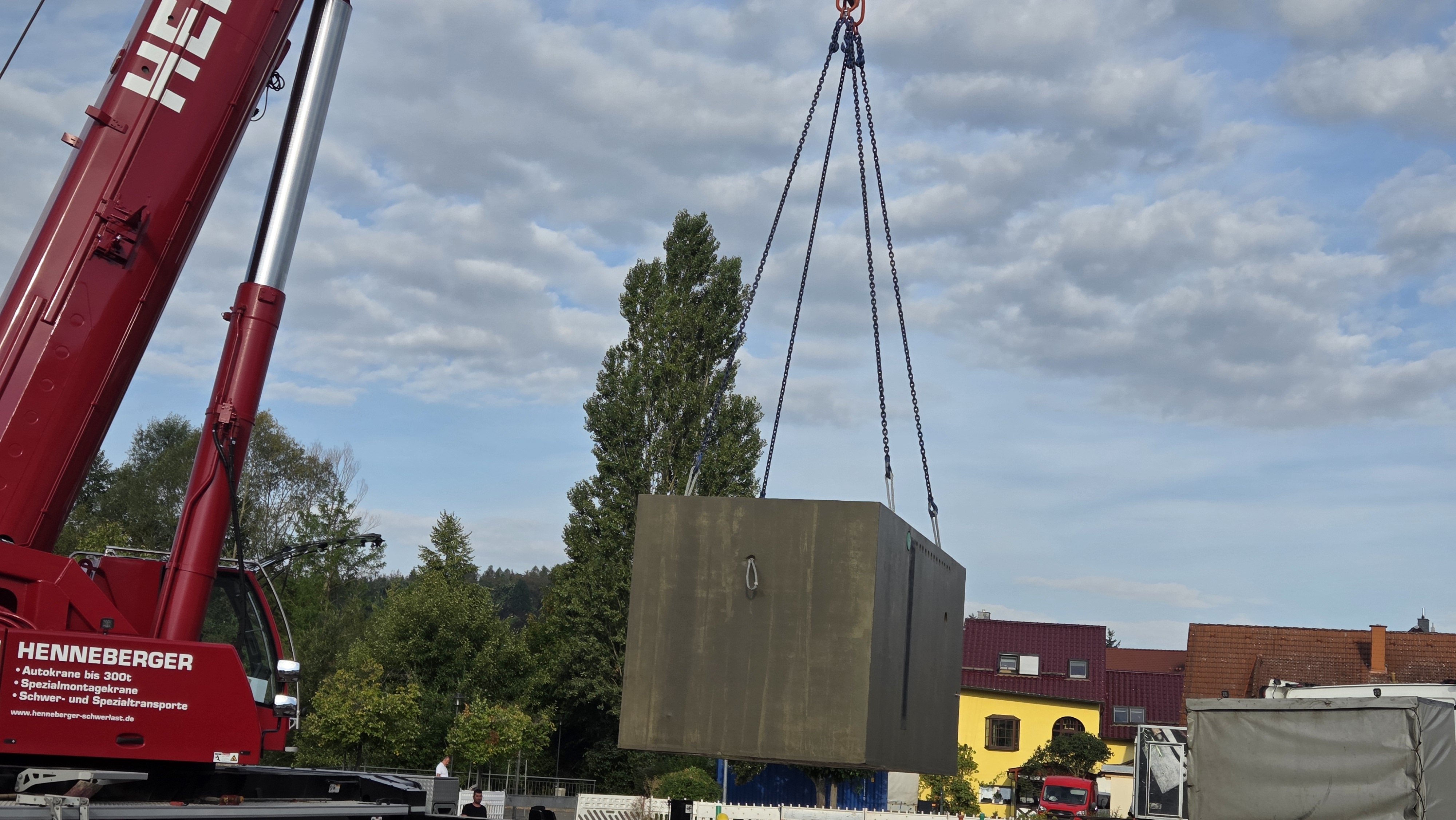 Brunnenkammer hängt an Kranaufhängung und schwebt zwischen Tieflader und Baugrube