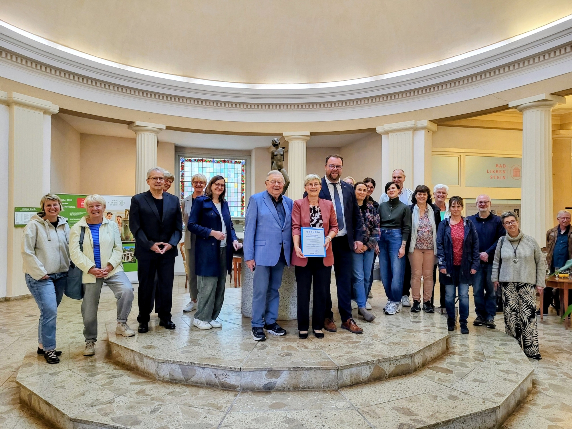 Gruppenbild ca. 20 Personen vor Brunnen in Wandelhalle, in der Mitte Bürgermeisterin und Staatssekretär mit Urkunde